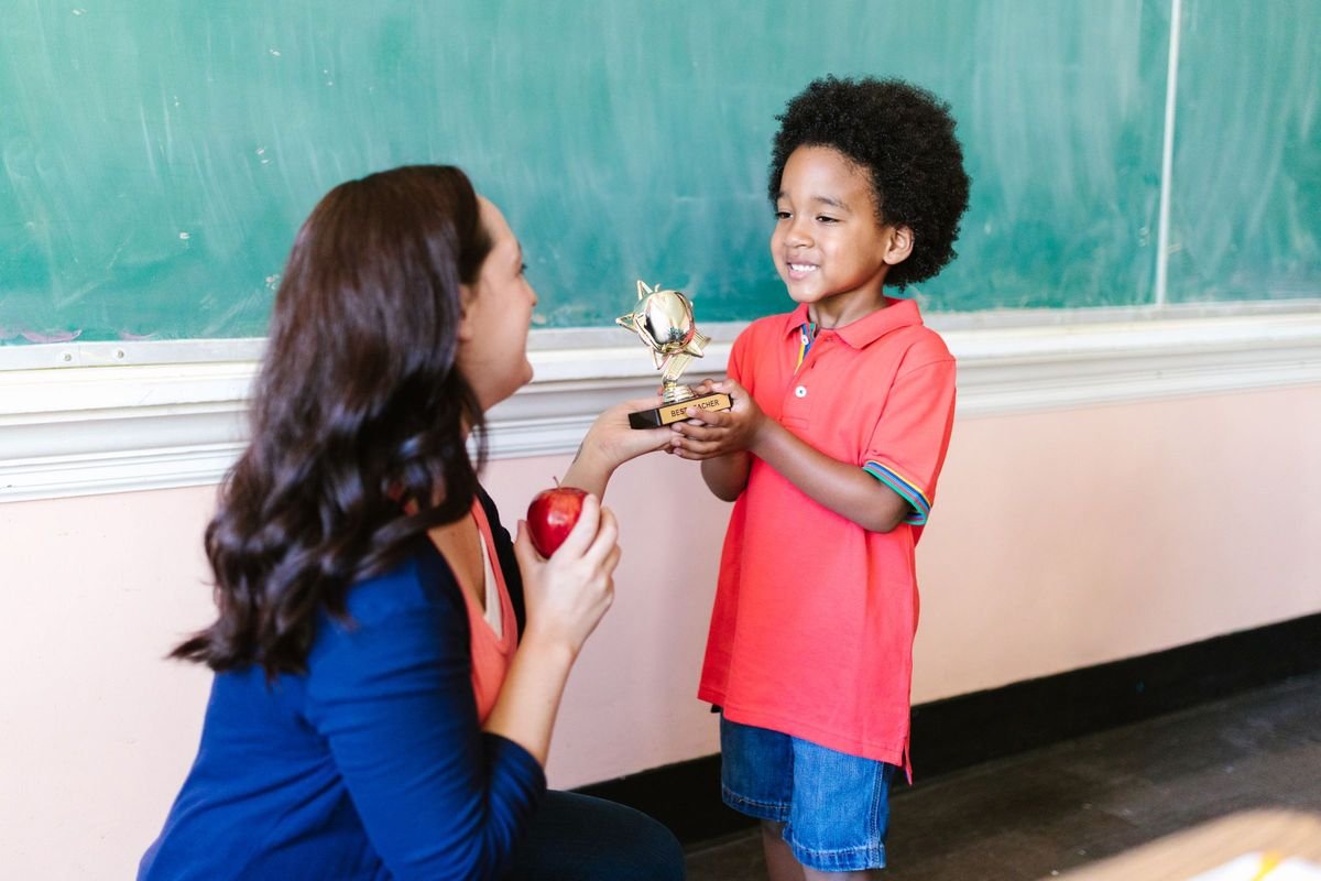 Teacher kneeling to celebrate with a preschooler holding a small gold trophy
